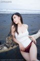 A woman in a white tank top sitting on a log on the beach.