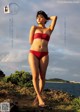 A woman in a red bikini standing on a rock by the water.